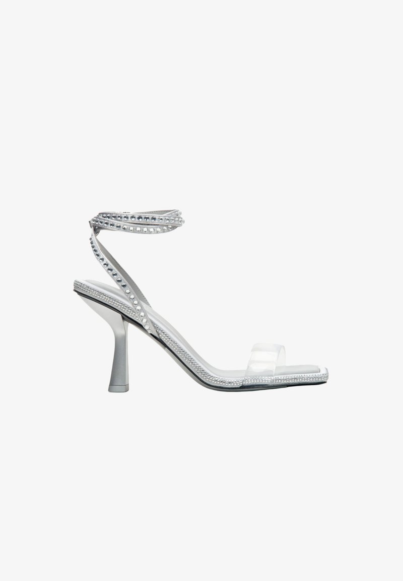 Przezroczyste sandały na obcasie z przezroczystym wierzchem, ozdobione srebrnymi ćwiekami i delikatnym paskiem na kostce, osadzone na smukłym obcasie.