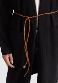 Personne portant un cardigan noir avec des poches frontales, noué à la taille avec une fine ceinture tressée marron, par-dessus une chemise blanche et un jean bleu.