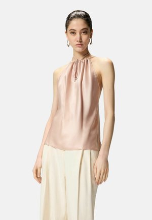 Donna con capelli scuri raccolti che indossa un top smanicato color rosa pallido a collo halter e pantaloni larghi color avorio, in piedi contro uno sfondo semplice.