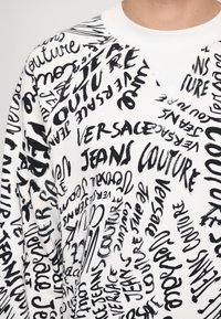 White sweatshirt featuring a graphic design of black handwritten text, including "Versace" and "Couture," with varying sizes and styles.