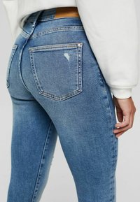 Jeans de mezclilla azul con un acabado desgastado, cinco bolsillos y un pequeño desgarro en el bolsillo trasero. Presenta una parche de cuero marrón en la cintura.
