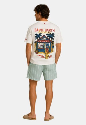 Uomo che indossa una maglietta bianca Saint Barth con illustrazione colorata di un negozio e una palma, pantaloncini a righe verdi e ciabatte beige, di spalle.
