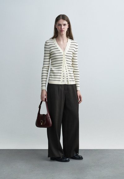 Γυναίκα που φοράει κρεμ και μαύρη ριγωτή ζακέτα με V-λαιμόκοψη, φαρδιά μαύρη παντελόνια, μαύρα παπούτσια, κρατώντας μπορντό τσάντα, στέκεται μπροστά σε απλό ανοικτό γκρι φόντο.