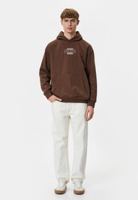Brun fleece hoodie med kængurulomme og grafisk tekst, parret med lyse jeans og hvide sneakers, stående mod en ensfarvet baggrund.