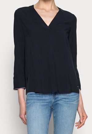 Kvinde iført en sort langærmet V-hals top kombineret med lyseblå jeans, stående mod en hvid baggrund.