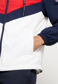 Color-blocked windbreaker in red, navy, and white. Features elastic cuffs, a zippered front, pockets, and an adjustable hem with toggle.