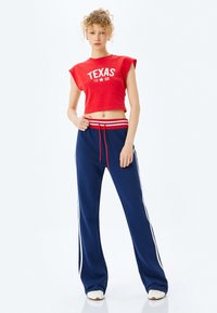 Rød crop sweatshirt med "TEXAS 1998" trykt i hvid, parret med navy sweatpants med hvide sidelister og en rød snor.