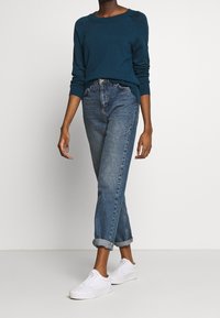 Maglione verde acqua, vestibilità ampia, colletto a giro, abbinato a jeans blu a vita alta, risvoltati alle caviglie, scarpe da ginnastica bianche. Outfit semplice e informale.