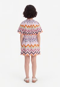 Niño con el pelo rizado de espaldas, vestido con un vestido de manga corta con patrón de zigzag multicolor y sandalias blancas sobre un fondo blanco.