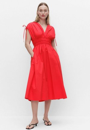 Kvinde iført en lyserød rød midi-kjole med rynket talje, korte ærmer med bånd og sorte hælede sandaler, står mod en hvid baggrund.