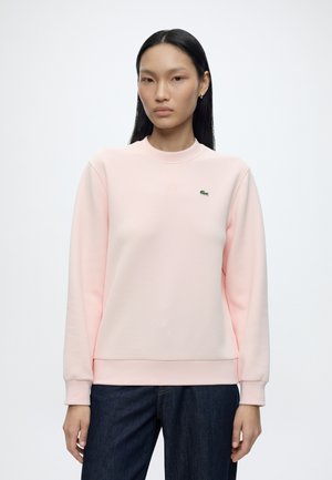 Femme aux longs cheveux noirs portant un sweat-shirt rose clair avec un petit logo crocodile vert sur la poitrine et un jean bleu foncé, debout devant un fond blanc.