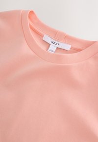 T-shirt rose doux à col rond avec étiquette de marque blanche cousue "NEXT" et étiquette de taille plus petite en dessous.