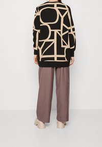 Osoba ubrana w czarny i beżowy sweter o abstrakcyjnym wzorze, luźne fioletowe spodnie i beżowe buty na platformie, stojąca na tle bez wzorów.