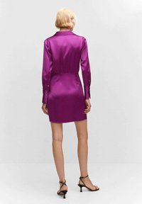 Frau mit kurzem blonden Haar, die ein glänzendes, lila Mini-Kleid mit langen Ärmeln und schwarze, geschnürte High-Heels trägt, steht vor einem weißen Hintergrund.