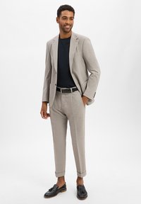 Lichtgrijs maatpak met een enkelvoudig jasje en geplooide broek, gecombineerd met een zwart shirt met ronde hals en zwarte instappers.