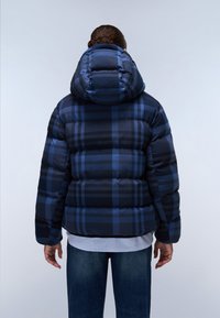Marineblå pufferjakke med plaidmønster, hætte og quiltet tekstur. Båret over en hvid skjorte og blå jeans. Bagfra.