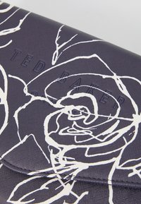 Granatowa kopertówka z białym nadrukiem róży, o teksturowanym materiale i wytłoczonym logo "TED BAKER" na przedniej klapie.