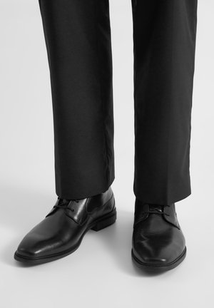 Czarne skórzane pantofle z gładkim wykończeniem i lekko teksturowaną częścią na palcach, zestawione z ciemnymi spodniami, prezentujące smukły design.