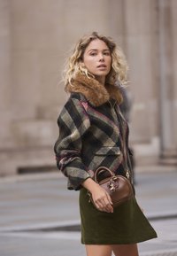 Wollkariertes Jackett mit braunem Pelzkragen, olivgrünem Rock und einer kleinen braunen Handtasche mit dekorativem Anhänger. Strukturiertes und geschichtetes Design.