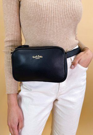 Femme portant un haut côtelé beige et un pantalon blanc avec une sacoche ceinture en cuir noir portant l'étiquette Gusti Leder studio autour de la taille.