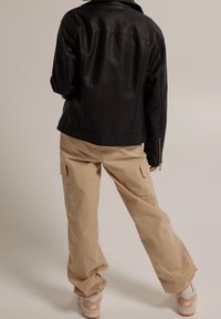 Giacca nera in ecopelle abbinata a pantaloni cargo beige larghi, con tasche laterali e cuciture a vista. Sneaker bianche con accenti rosa.