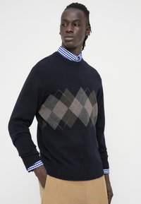 Vīrietis, kurš valkā tumši zilu džemperi ar pelēku un brūnu argaila rakstu, zem tā zilā rūtainā krekla, baltas bikses, rokas kabatās, skatās pa kreisi.