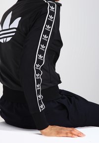 Černá sportovní bunda s bílými pruhy a logy po stranách rukávů, žebrované manžety a límec. Hladká tkanina, přiléhavý design s výrazným odznakem.