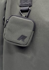 Donkergroene stoffen tas met een kleine ritszak, voorzien van een zwart logo, een textuur oppervlak en een metalen clip bevestiging.