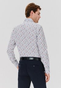 Męska koszula z długim rękawem w białym kolorze z kwiatowym wzorem w odcieniach niebieskiego i czerwonego. Posiada kołnierzyk zapinany na guziki oraz Slim Fit, zestawiona z ciemnymi spodniami.