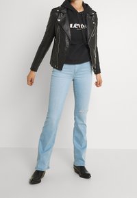 Czarna skórzana kurtka motocyklowa na czarnej bluzie z kapturem z logo, w zestawieniu z jasnoniebieskimi dżinsami w stylu dzwonów i czarnymi botkami na kostkę.