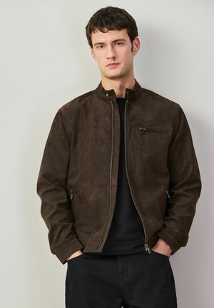 Junger Mann trägt eine dunkelbraune Jacke mit Reißverschluss, Stehkragen und Reißverschlusstasche auf der Brust, steht vor einer schlichten hellen Wand.