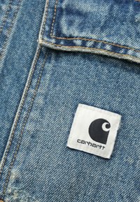 Υλικό ντένιμ σε ανοιχτό μπλε με ελαφρώς ξεθωριασμένη υφή. Διαθέτει αντίθετες πορτοκαλί ραφές και μια λευκή υφασμάτινη ετικέτα με μαύρο λογότυπο.