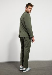 Oliven grøn jakkesæt med en skræddersyet blazer og matchende bukser. Båret med hvide og grønne sneakers. Glat stof, moderne snit og minimal detaljer.