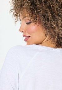 Top de tela blanca y ligera con cuello redondo y mangas raglán; cabello rizado visible de perfil, mostrando la textura y el color.