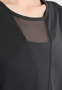 Top negro con diseño de cuello en V, paneles de malla y tejido texturizado, que muestra una combinación de superficies suaves y perforadas.