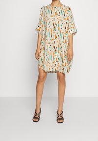 Robe à manches courtes, longueur genou, de couleur beige clair avec un motif abstrait coloré composé de formes orange, verte et noire. Tissu doux ; coupe décontractée.