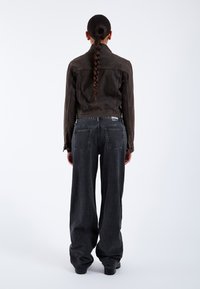 Person med langt flet hår iført en mørk kort jakke og løstsiddende sorte jeans, stående med ryggen mod en ensfarvet hvid baggrund.