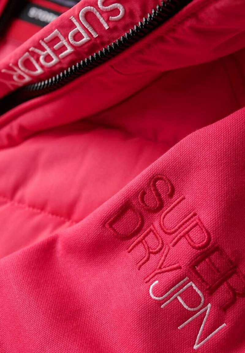 Rote Pufferjacke mit glatter Textur und sichtbaren Nähten. Mit gesticktem Text "SUPER DRY" in Weiß und dunklerem Rot. Reißverschlussdetails.
