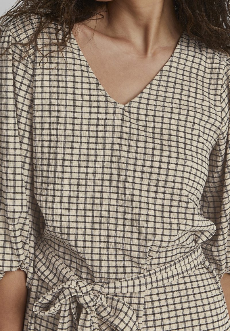 Femme portant une robe à carreaux beige et noire avec un col en V et une ceinture nouée à la taille, montrée des épaules jusqu'aux hanches.