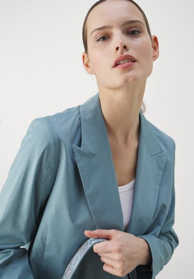 Blazer azzurro chiaro con colletto a revers, tessuto testurizzato e vestibilità rilassata. In mano è tenuto un cinturino regolabile a fantasia con accenti blu.