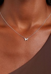 Collar de mariposa de plata con una cadena delicada, que presenta un colgante de mariposa adornado con pequeñas piedras de strass, descansando sobre una tela marrón.