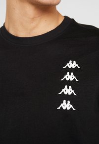 Černé bavlněné tričko s kulatým výstřihem, se čtyřmi bílými potisky loga v svislém uspořádání na levé straně hrudi.