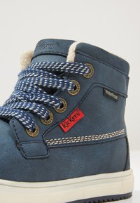 Bota impermeable azul con cordones redondos, ojales de metal y parche de logo rojo. Presenta un upper texturizado y costuras blancas en la suela.