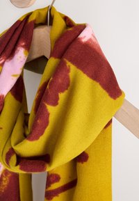 Kootud sall, millel on kollane alus ja burgundia ning roosa batikmustrid. Pehme tekstuur; lahtiselt puidust riidepuul rippumas.