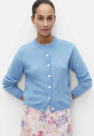 Kobieta z włosami zebranymi do tyłu, nosząca kolczyki koła, jasnoniebieski rozpinany kardigan i pastelową kwiecistą spódnicę, stojąca na białym tle.