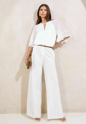 Weiße Bluse mit transparenten, schwingenden Ärmeln und einem Schlüsseloch-Ausschnitt, kombiniert mit weißen weit geschnittenen Hosen und einem braunen Gürtel, hält eine helle Clutch.