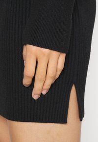 Abito nero a coste con uno spacco laterale, caratterizzato da un motivo in maglia testurizzato. Una mano è posizionata sul tessuto, enfatizzando la sua morbidezza e i dettagli.
