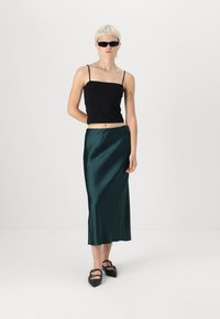 Crna majica bez rukava, tamnozelena saten midi suknja i crne cipele s visokim potpeticama s detaljima vezivanja. Model ima kratku platinastu kosu i naočale za sunce.