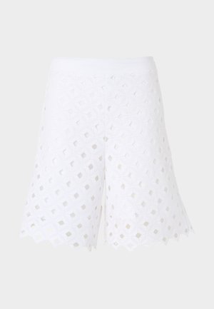 Frau trägt ärmelloses weißes Top und passende Shorts aus Lochspitze, hält schwarze und weiße geflochtene Korbtasche, schwarze Sandalen, steht.