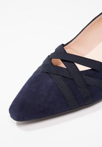 Chaussure plate en daim bleu marine avec un bout pointu, équipée de brides élastiques croisées sur la tige et d'une doublure intérieure lisse.